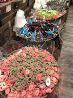 A display of various colorful candies in large bowls lined with cellophane wrappers. The nearest bowl contains pink and green gummy candies shaped like rings and strawberries. The middle bowl features an assortment of blue gummy dolphins mixed with red and black gummies. The far bowl contains green and red candies shaped like watermelon slices. Clear plastic bags for filling candy are hanging on a rustic brick and wooden backdrop.