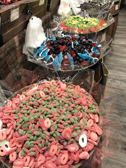 A display of various colorful candies in large bowls lined with cellophane wrappers. The nearest bowl contains pink and green gummy candies shaped like rings and strawberries. The middle bowl features an assortment of blue gummy dolphins mixed with red and black gummies. The far bowl contains green and red candies shaped like watermelon slices. Clear plastic bags for filling candy are hanging on a rustic brick and wooden backdrop.