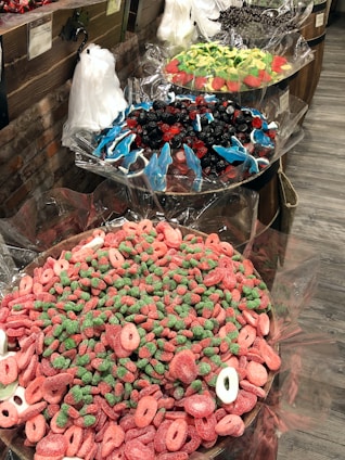 A display of various colorful candies in large bowls lined with cellophane wrappers. The nearest bowl contains pink and green gummy candies shaped like rings and strawberries. The middle bowl features an assortment of blue gummy dolphins mixed with red and black gummies. The far bowl contains green and red candies shaped like watermelon slices. Clear plastic bags for filling candy are hanging on a rustic brick and wooden backdrop.