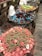 A display of various colorful candies in large bowls lined with cellophane wrappers. The nearest bowl contains pink and green gummy candies shaped like rings and strawberries. The middle bowl features an assortment of blue gummy dolphins mixed with red and black gummies. The far bowl contains green and red candies shaped like watermelon slices. Clear plastic bags for filling candy are hanging on a rustic brick and wooden backdrop.