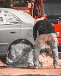 A person is working on a car, likely engaged in some form of maintenance or repair. The car's wheel area is covered, and there is a hose or cable nearby. The orange vehicle or truck is parked closely in the background.