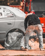 A person is working on a car, likely engaged in some form of maintenance or repair. The car's wheel area is covered, and there is a hose or cable nearby. The orange vehicle or truck is parked closely in the background.