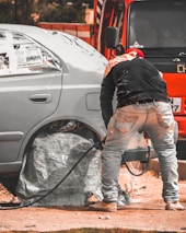 A person is working on a car, likely engaged in some form of maintenance or repair. The car's wheel area is covered, and there is a hose or cable nearby. The orange vehicle or truck is parked closely in the background.