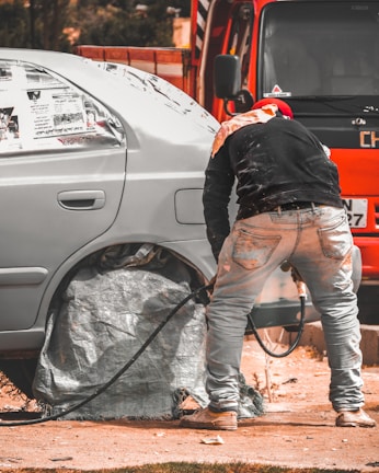 A person is working on a car, likely engaged in some form of maintenance or repair. The car's wheel area is covered, and there is a hose or cable nearby. The orange vehicle or truck is parked closely in the background.