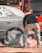 A person is working on a car, likely engaged in some form of maintenance or repair. The car's wheel area is covered, and there is a hose or cable nearby. The orange vehicle or truck is parked closely in the background.