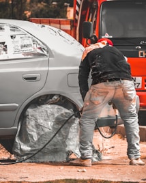 A person is working on a car, likely engaged in some form of maintenance or repair. The car's wheel area is covered, and there is a hose or cable nearby. The orange vehicle or truck is parked closely in the background.
