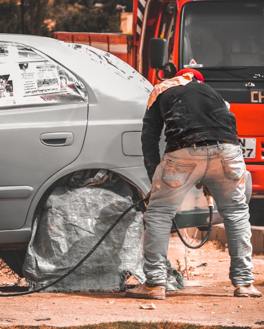 A person is working on a car, likely engaged in some form of maintenance or repair. The car's wheel area is covered, and there is a hose or cable nearby. The orange vehicle or truck is parked closely in the background.