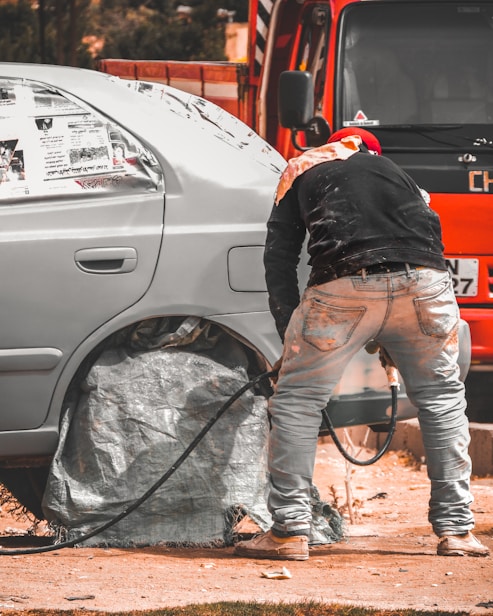 A person is working on a car, likely engaged in some form of maintenance or repair. The car's wheel area is covered, and there is a hose or cable nearby. The orange vehicle or truck is parked closely in the background.