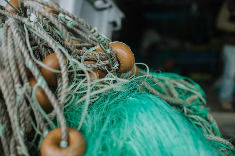 A close-up of a fishing net with brown floats attached. The net is made of thick green and beige fibers and appears to be coiled or piled up, suggesting it's not currently in use. The background is out of focus, with indistinct dark and blurred forms.