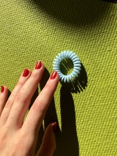A hand with red-painted nails reaches toward a blue coiled hair tie placed on a green textured surface.
