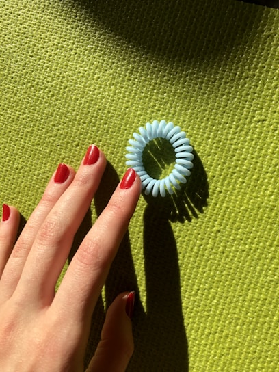 A hand with red-painted nails reaches toward a blue coiled hair tie placed on a green textured surface.
