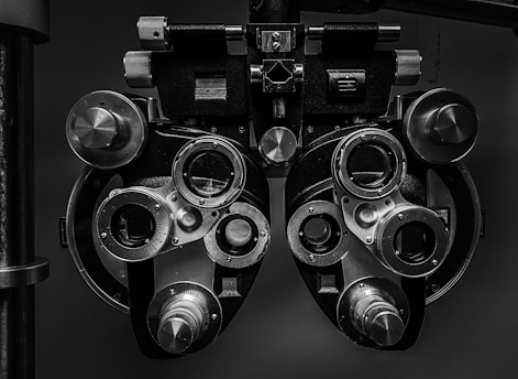 A close-up view of an optometry device called a phoropter, used for testing individual lenses on each eye during an eye exam. The device is composed of multiple dials, lenses, and knobs.