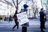 A group of people participate in a protest on a city street. One individual is prominently holding a sign that reads 'ACT NOW or SWIM later.' They are dressed warmly, suggesting a cooler climate, and are wearing face masks. The background features leafless trees and buildings.