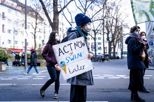 A group of people participate in a protest on a city street. One individual is prominently holding a sign that reads 'ACT NOW or SWIM later.' They are dressed warmly, suggesting a cooler climate, and are wearing face masks. The background features leafless trees and buildings.