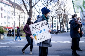 A group of people participate in a protest on a city street. One individual is prominently holding a sign that reads 'ACT NOW or SWIM later.' They are dressed warmly, suggesting a cooler climate, and are wearing face masks. The background features leafless trees and buildings.