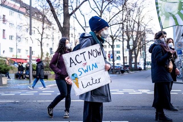 A group of people participate in a protest on a city street. One individual is prominently holding a sign that reads 'ACT NOW or SWIM later.' They are dressed warmly, suggesting a cooler climate, and are wearing face masks. The background features leafless trees and buildings.