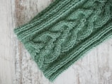 A close-up of green knitted fabric featuring a detailed cable knit pattern. The soft texture contrasts with the rustic wooden surface beneath.