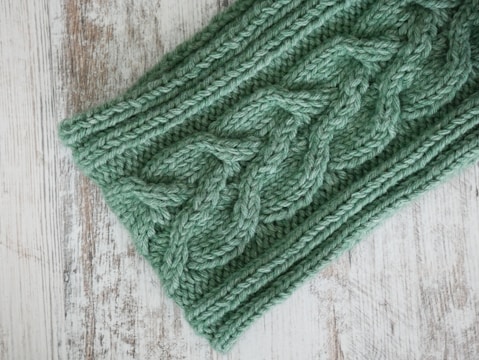 A close-up of green knitted fabric featuring a detailed cable knit pattern. The soft texture contrasts with the rustic wooden surface beneath.