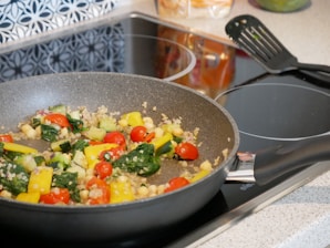 A frying pan on an electric stove contains a mix of colorful vegetables including red cherry tomatoes, yellow bell peppers, green spinach, zucchini, and small grains such as quinoa. A black spatula lies on the stove beside the pan. The background includes a patterned backsplash and some kitchen items.