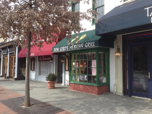A street view of several storefronts including a Mexican grill with a green awning, a tree in front of the shops with brown leaves, and a sidewalk lined with bricks.