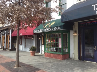 A street view of several storefronts including a Mexican grill with a green awning, a tree in front of the shops with brown leaves, and a sidewalk lined with bricks.