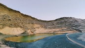 A serene lake nestled within a rocky, arid landscape. The surrounding rock formations exhibit layered textures and earthy tones, while the calm water of the lake reflects hues of green and yellow. The sky overhead is clear and vast, with a faint hint of blue. There is also a small, winding path bordered by a low fence that follows the contour of the lake and leads off into the distance.
