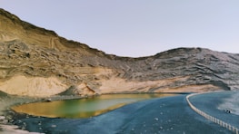 A serene lake nestled within a rocky, arid landscape. The surrounding rock formations exhibit layered textures and earthy tones, while the calm water of the lake reflects hues of green and yellow. The sky overhead is clear and vast, with a faint hint of blue. There is also a small, winding path bordered by a low fence that follows the contour of the lake and leads off into the distance.