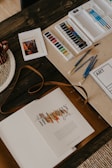 An arrangement of art materials including watercolors, paint tubes, and various paintbrushes is displayed on a wooden table. A book titled 'The History Book' lies open, alongside a spiral-bound sketchbook labeled 'ART'. A woven placemat and a striped mug are partially visible.