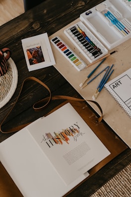 An arrangement of art materials including watercolors, paint tubes, and various paintbrushes is displayed on a wooden table. A book titled 'The History Book' lies open, alongside a spiral-bound sketchbook labeled 'ART'. A woven placemat and a striped mug are partially visible.