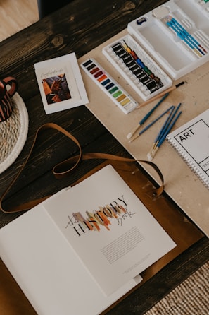 An arrangement of art materials including watercolors, paint tubes, and various paintbrushes is displayed on a wooden table. A book titled 'The History Book' lies open, alongside a spiral-bound sketchbook labeled 'ART'. A woven placemat and a striped mug are partially visible.