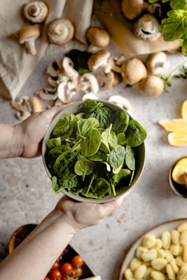 Hands holding a bowl of fresh spinach leaves, surrounded by various food items including whole and sliced mushrooms, cherry tomatoes, gnocchi, sliced lemons, and a beige cloth.
