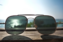 A pair of sunglasses is positioned upright on a wooden surface with a blurred background of the ocean and a clear sky. The sunlight reflects off the sea, creating a bokeh effect visible through the tinted lenses.