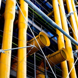 Large yellow industrial pipes are prominently featured, supported by a framework of metal rods and wires. The structure appears to be part of a modern building facade with a mix of exposed pipes and metalwork.