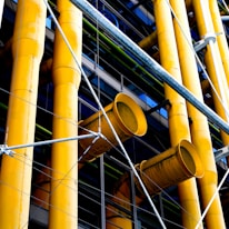 Large yellow industrial pipes are prominently featured, supported by a framework of metal rods and wires. The structure appears to be part of a modern building facade with a mix of exposed pipes and metalwork.