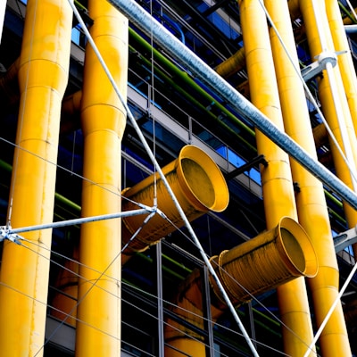 Large yellow industrial pipes are prominently featured, supported by a framework of metal rods and wires. The structure appears to be part of a modern building facade with a mix of exposed pipes and metalwork.