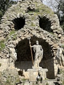 A stone grotto features a prominent statue of a mythological figure holding a trident. The figure stands surrounded by other carved figures in flowing garments. The stone backdrop resembles a natural rock formation with vegetation interspersed.