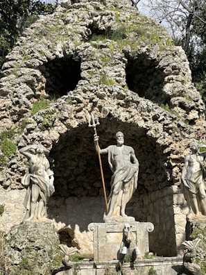 A stone grotto features a prominent statue of a mythological figure holding a trident. The figure stands surrounded by other carved figures in flowing garments. The stone backdrop resembles a natural rock formation with vegetation interspersed.