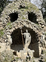 A stone grotto features a prominent statue of a mythological figure holding a trident. The figure stands surrounded by other carved figures in flowing garments. The stone backdrop resembles a natural rock formation with vegetation interspersed.