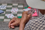 An elderly person holding a hand of playing cards near a chessboard. The focus is on the person's hands showing signs of age and a partial view of their face obscured by a hat. The surface is speckled and there are scattered playing cards and poker chips in the background.