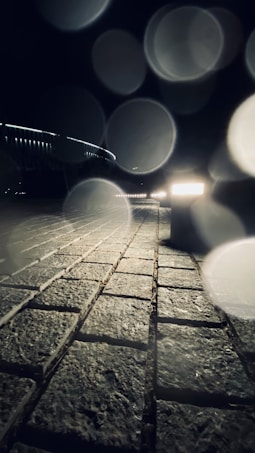 A nighttime scene with a brick pathway in the foreground leading into the distance. The path is illuminated by lights, with soft, circular light artifacts visible across the image. A railing on the left side of the path suggests it might be a bridge or walkway.