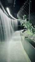 A modern, dimly-lit corridor features a unique water installation with cascading waterfalls on one side and lush green vines growing along a wired trellis on the other. The walkway is bordered by sleek, black stone planters filled with gravel. Overhead, linear light fixtures provide subtle illumination, creating a serene and futuristic atmosphere.