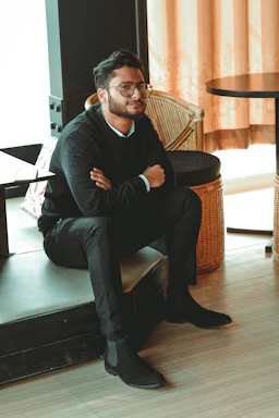 A man wearing glasses and dressed in business casual attire is seated with his arms crossed. He is sitting on the edge of a platform or raised surface in a room with bamboo furniture and light-colored curtains. The setting appears to be modern and cozy, with a blend of contemporary and natural elements.