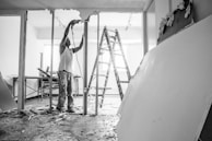 A person wearing work clothes and standing on a ladder is involved in construction or renovation. Debris is scattered on the floor, and large drywall pieces are leaning against the wall. There is a table in the background near a window, and the atmosphere suggests active construction work.