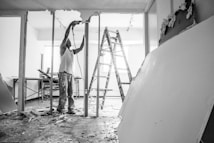 A person wearing work clothes and standing on a ladder is involved in construction or renovation. Debris is scattered on the floor, and large drywall pieces are leaning against the wall. There is a table in the background near a window, and the atmosphere suggests active construction work.