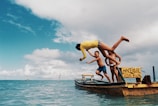 Children joyfully jump off a wooden boat into clear blue water under a vibrant sky. The scene exudes a carefree summertime atmosphere with clouds scattered across the expanse of the sky.