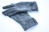 A pair of gray woolen gloves is scattered with small white snowflakes, resting on a smooth white surface. The gloves have a simple design with three small buttons on one side and a seam running around the wrist area.
