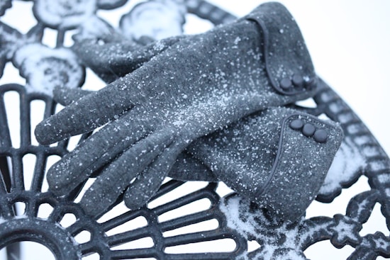 A pair of dark gray gloves are resting on a metal outdoor chair. The gloves are dusted with a light layer of snow, enhancing the wintery atmosphere. The chair features an intricate pattern, and traces of snow can be seen on the chair surface as well.