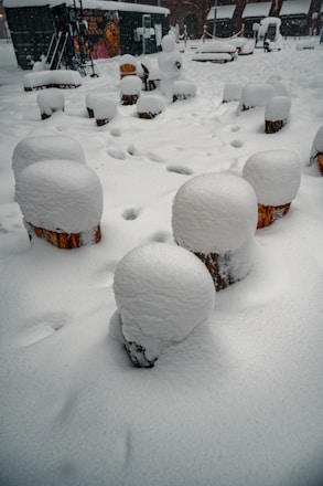 A snowy outdoor scene with logs covered in thick layers of snow, arranged in a circle. A snowman can be seen in the center background, with snow continuing to fall, creating a wintry atmosphere.