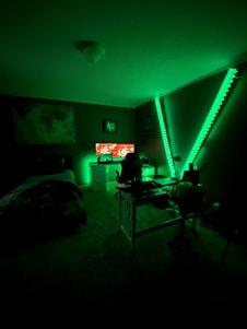A dimly lit room with green lighting features a bed, a person sitting at a desk with two computer monitors displaying vibrant red screens, and LED strip lights creating a V-shape on the wall.