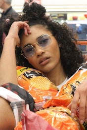 A person with curly hair and wearing sunglasses is surrounded by bags of potato chips. They are resting their head on one hand and appear relaxed. The setting seems to be a casual environment, possibly a store.